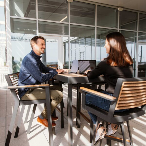 Man and woman smiling while collaborating on laptops on an outdoor rooftop workspace.