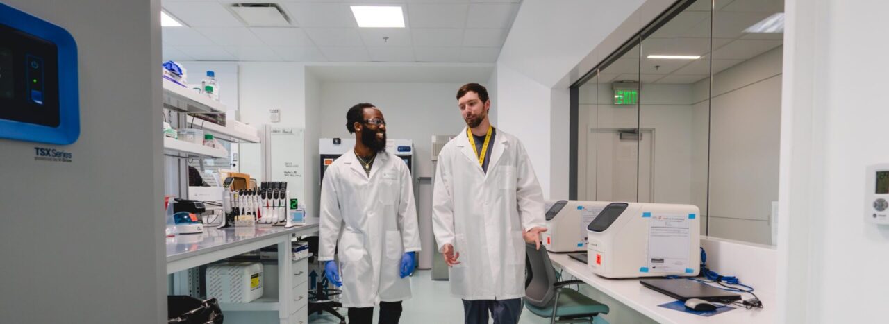 Two scientists in lab coats walking and talking through a research laboratory.