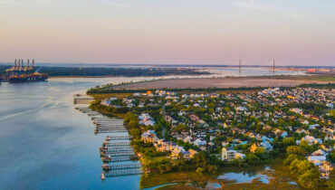 Aerial view of Charleston's waterfront featuring residential neighborhoods, marshlands, the Wando River, cargo ships at the Port of Charleston, and the Arthur Ravenel Jr. Bridge in the distance.