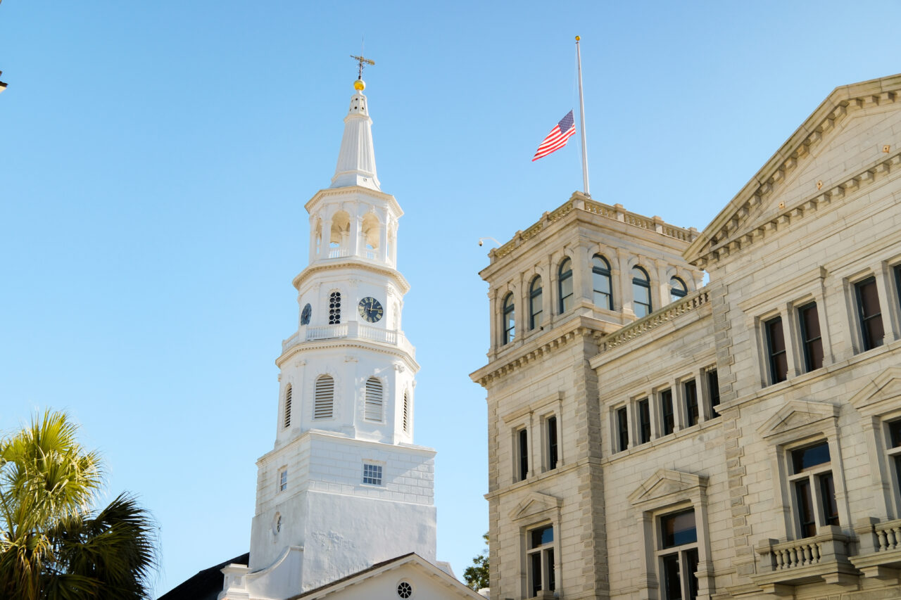 White church steeple next to a historic stone building flying the American flag in Charleston.