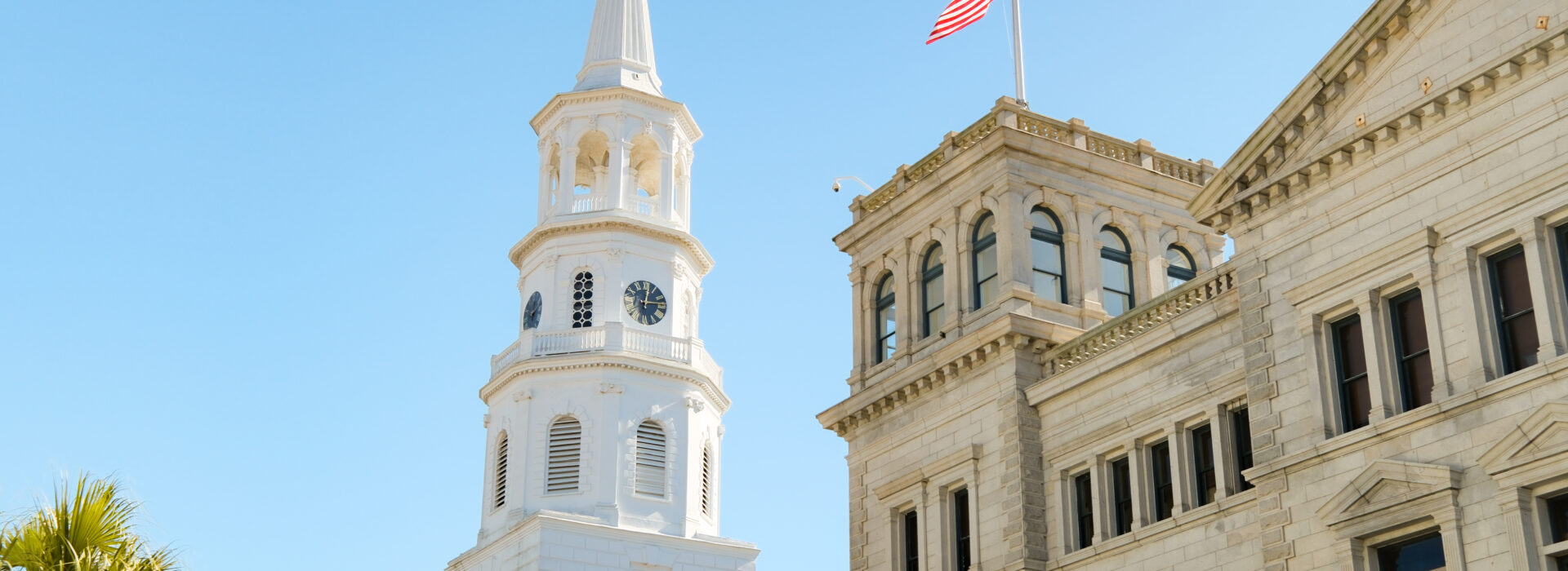 White church steeple next to a historic stone building flying the American flag in Charleston.