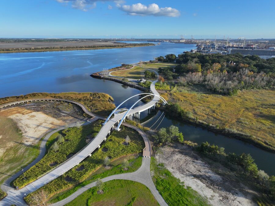 Aerial view of a riverside park with a curved pedestrian bridge and walking trails.