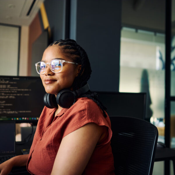 young BIPOC woman sits in front of computers, looking at camera