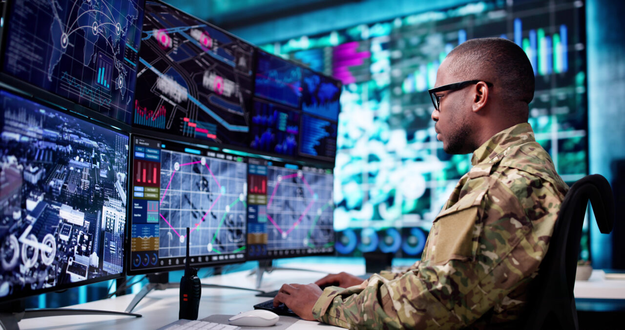 Military service member working at a computer workstation with multiple digital screens displaying maps, data, and cybersecurity information.