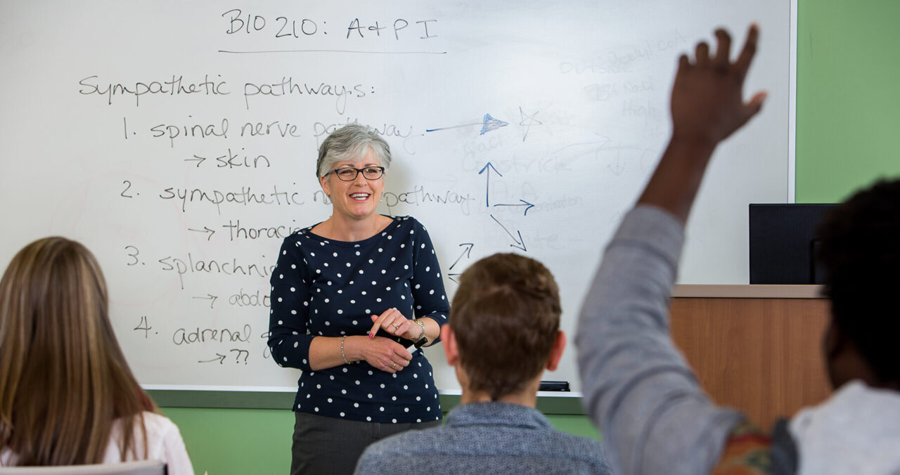 A science teacher in front of a whiteboard in a classroom sees a student raise his hand.