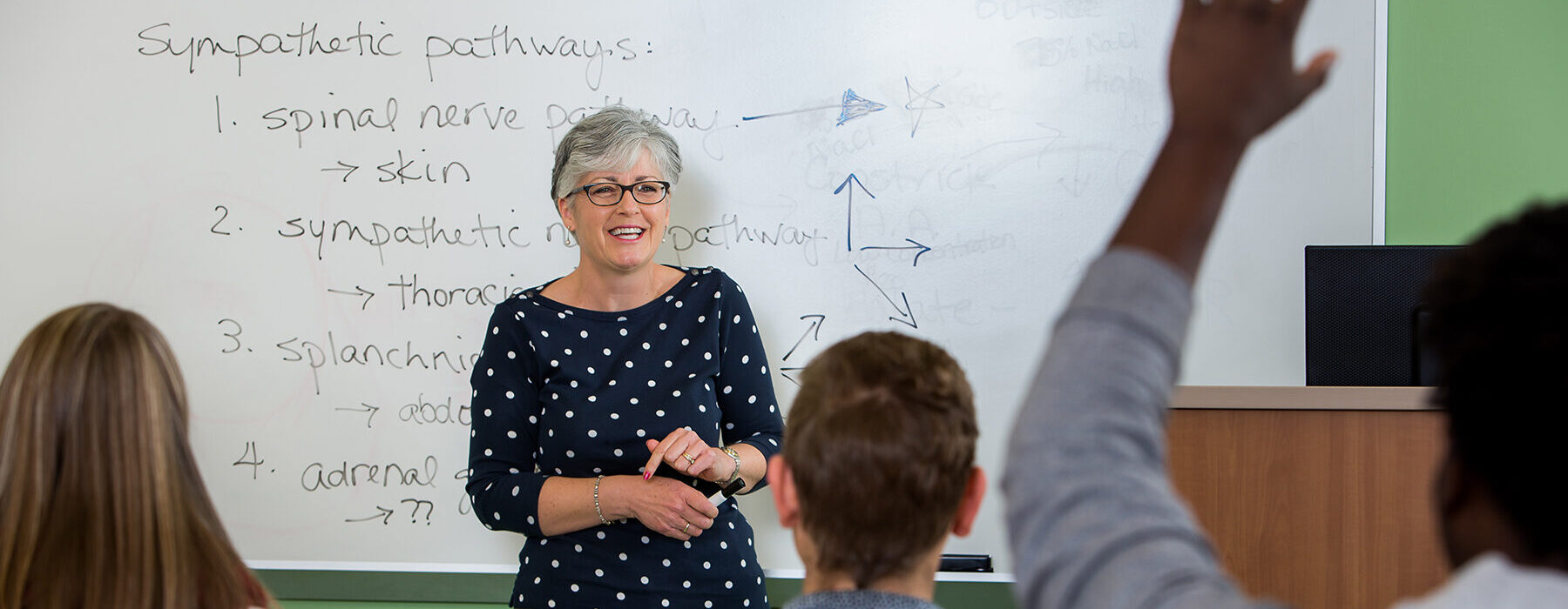 A science teacher in front of a whiteboard in a classroom sees a student raise his hand.