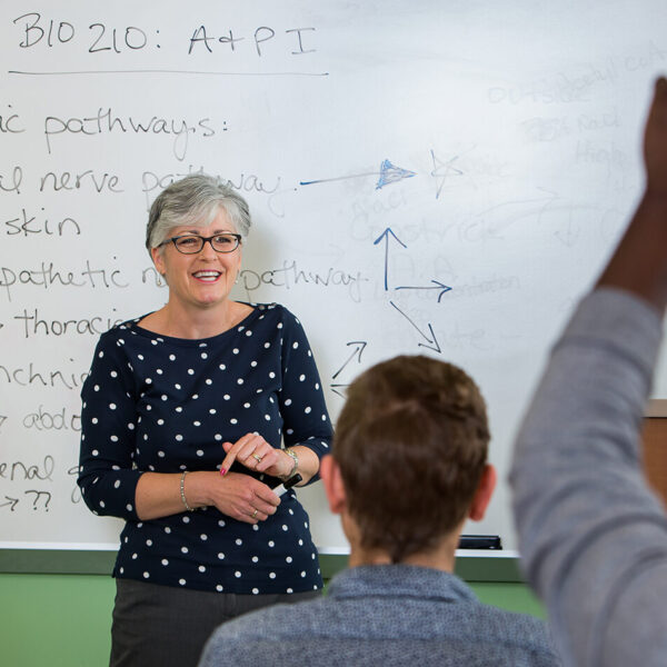 A science teacher in front of a whiteboard in a classroom sees a student raise his hand.