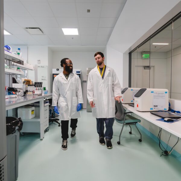 Two young men in lab coats walk through a lab together.