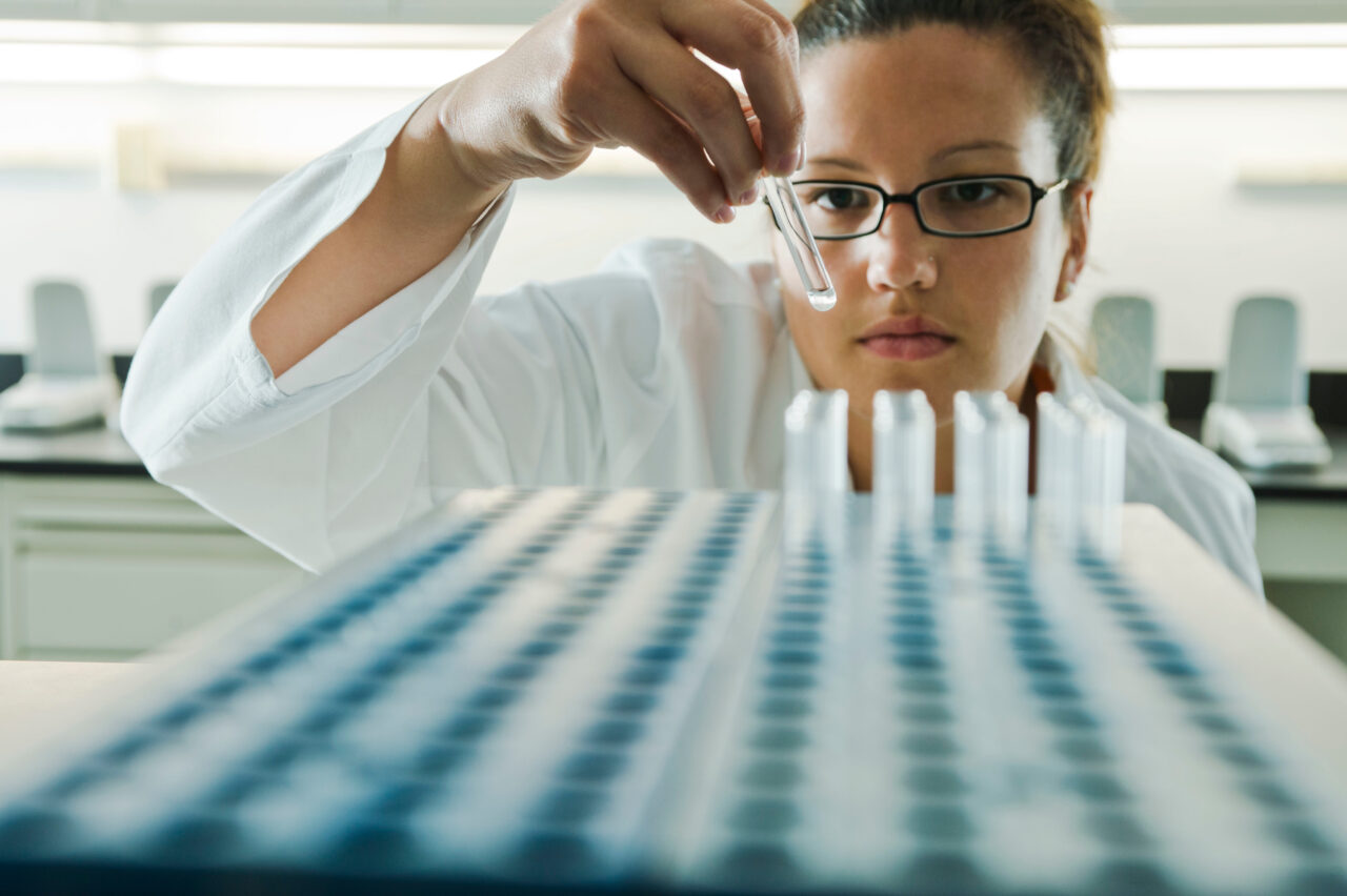 A scientist performs a quality assurance inspection on a series of test tubes.