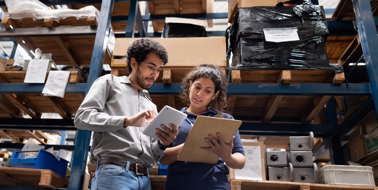 Two coworkers review an iPad and a clipboard together in a fulfillment warehouse.