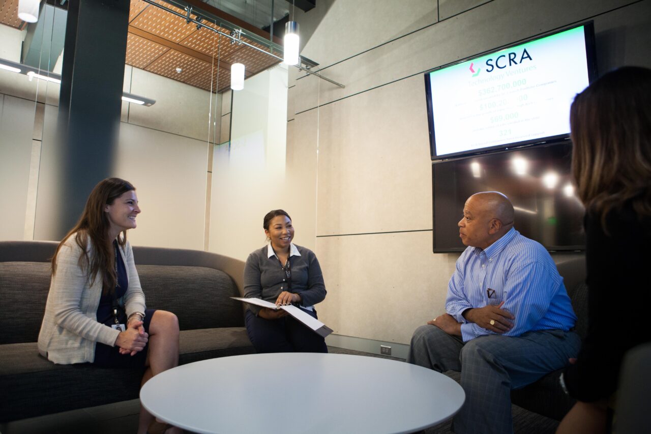 Three business people sit around a casual office table and talk together.